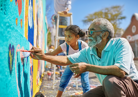 Community members work together to paint a colorful mural in a welcoming outdoor setting, creating an upbeat and inclusive atmosphere